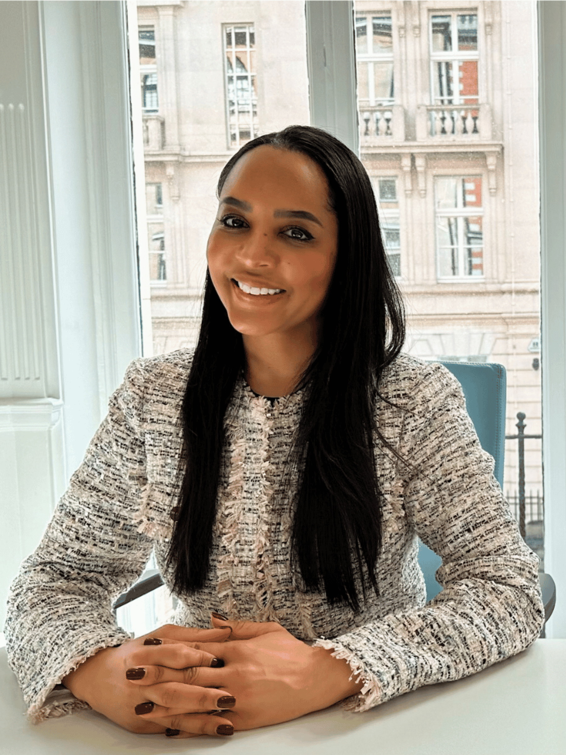 Ms. Natasha Berridge, a smiling woman with long dark hair, sitting behind a desk wearing a light tweed jacket, with her hands crossed, wearing dark nail varnish